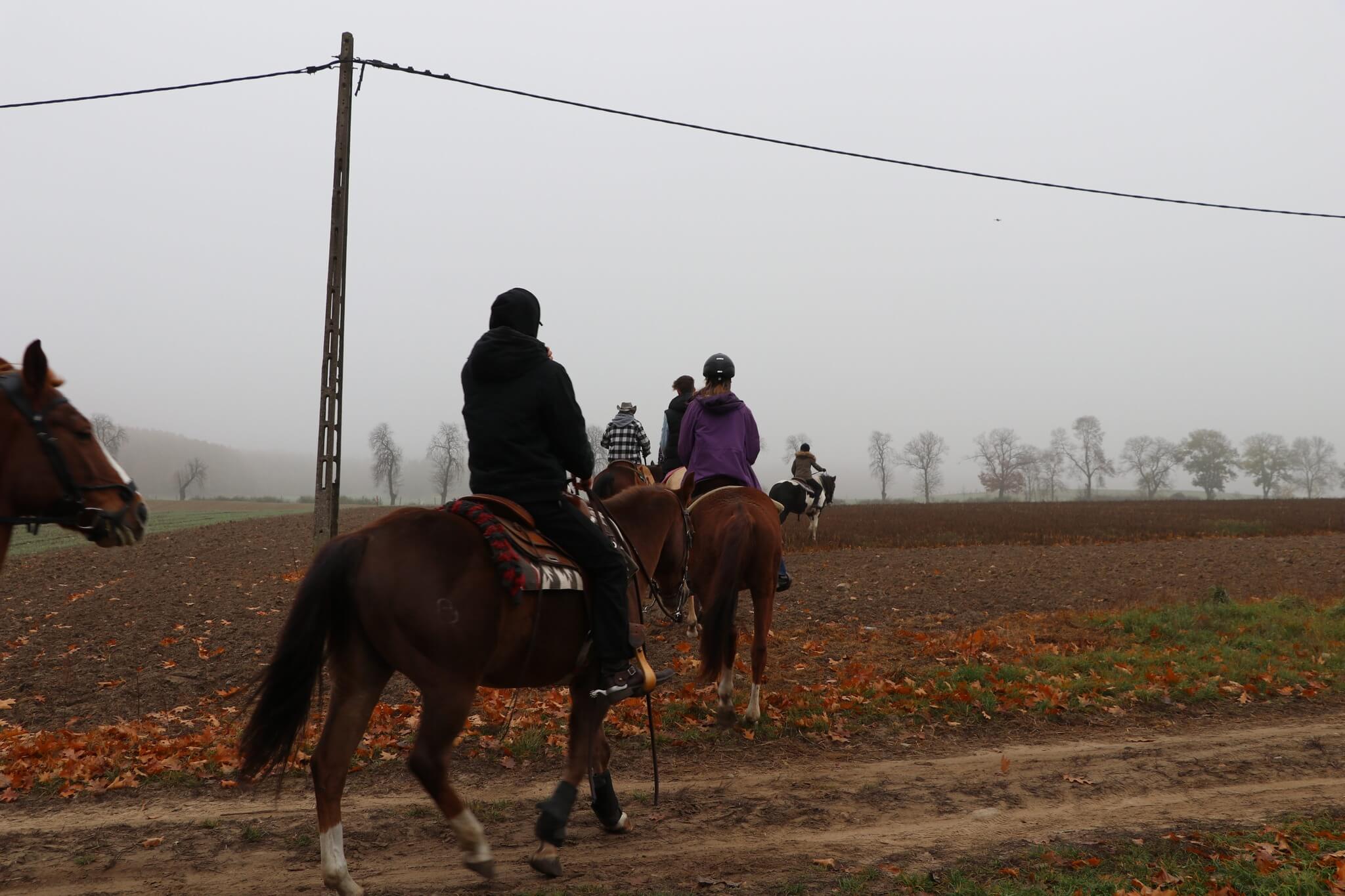 Grupa jeźdźców jadąca gęsiego polną drogą w jesiennej mgle